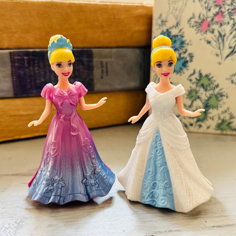 May include: Two Cinderella dolls. One doll wears a pink and blue ombre gown with a bow. The other doll wears a white gown with a blue underskirt. Both dolls have blonde hair and blue headbands.