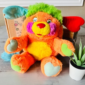 May include: A plush toy with orange fur, a bright green tuft of hair on top, and a yellow belly. It features one teal ear, one red ear, purple cheek patches, a red nose, and light green heart-shaped accents on the paws and feet.