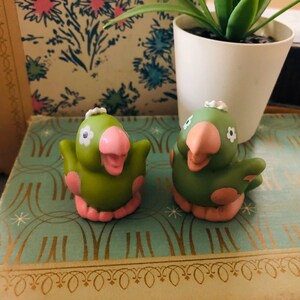 Pu&ograve; includere: Due statuette di pappagalli verdi con becchi, zampe e dettagli rosa. Ogni uccello ha un fiore bianco sulla testa ed &egrave; posato con le ali spiegate. Le statuette sono su una superficie decorativa.