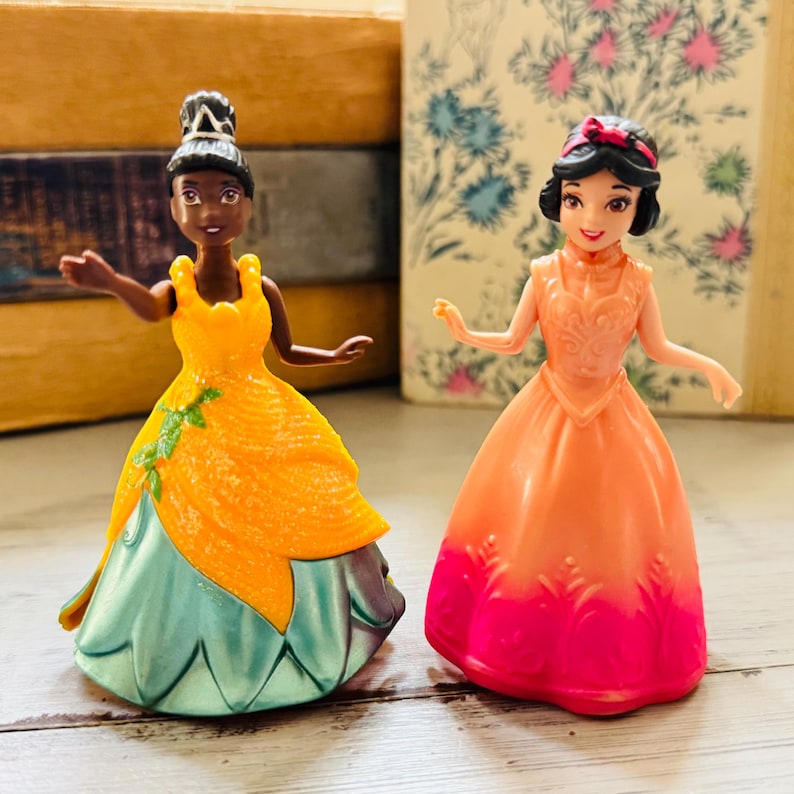 May include: Two small princess figurines. One wears a yellow and teal dress, the other an orange and pink dress. Both have dark hair and are posed with their arms outstretched. The figurines are on a light-colored surface.