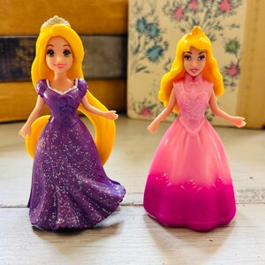 May include: Two small princess dolls. One doll wears a purple dress with glitter and a tiara. The other doll wears a pink dress with a pink and purple ombre effect. Both dolls have yellow hair.
