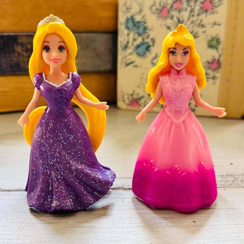 May include: Two small princess dolls. One doll wears a purple dress with glitter and a tiara. The other doll wears a pink dress with a pink and purple ombre effect. Both dolls have yellow hair.