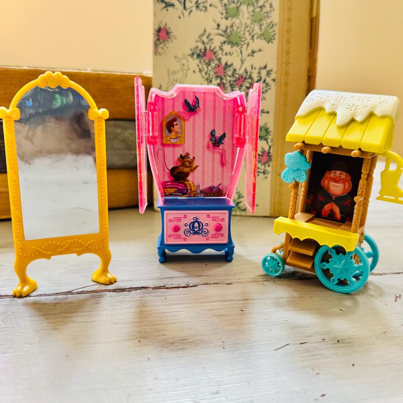 May include: A yellow decorative mirror, a pink and blue miniature cabinet with Cinderella figures, and a yellow and blue toy wagon with a character inside. The toys are on a light-colored surface.