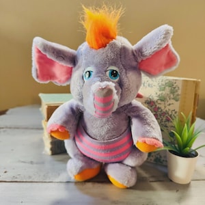 Op de afbeelding: Een pluchen olifant in grijstinten, roze en oranje. De olifant heeft grote oren met roze binnenkanten, blauwe ogen en een gestreepte slurf. Hij heeft oranje haar en een roze en grijs gestreepte buikband. Het speelgoed staat rechtop.