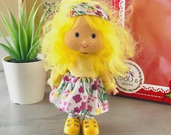 May include: A vintage doll with yellow hair, a yellow top, and a floral skirt. The doll has freckles and is wearing a floral headband and yellow shoes. The packaging in the background has the text "1980s Design".