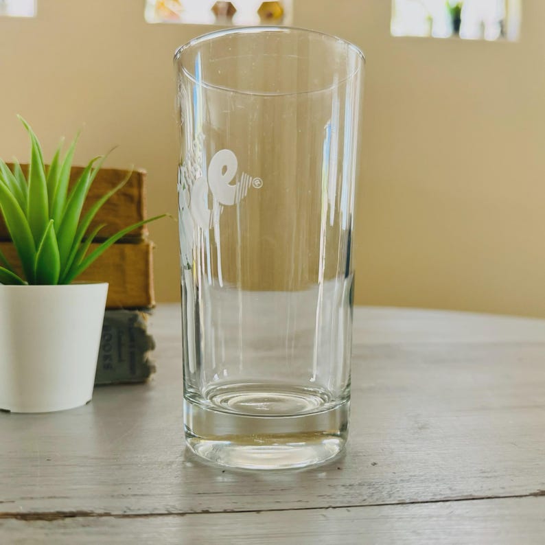 Pu&ograve; includere: Un bicchiere cilindrico trasparente con un logo bianco sul lato. Il bicchiere &egrave; vuoto e si trova su una superficie chiara. Una piccola pianta in vaso &egrave; visibile a sinistra del bicchiere.
