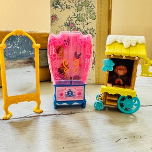 May include: A yellow toy mirror, a pink and blue toy wardrobe, and a yellow and blue toy cart are arranged on a light-colored surface. The mirror has a decorative frame. The wardrobe has a pink frame and blue base. The cart has a character inside.