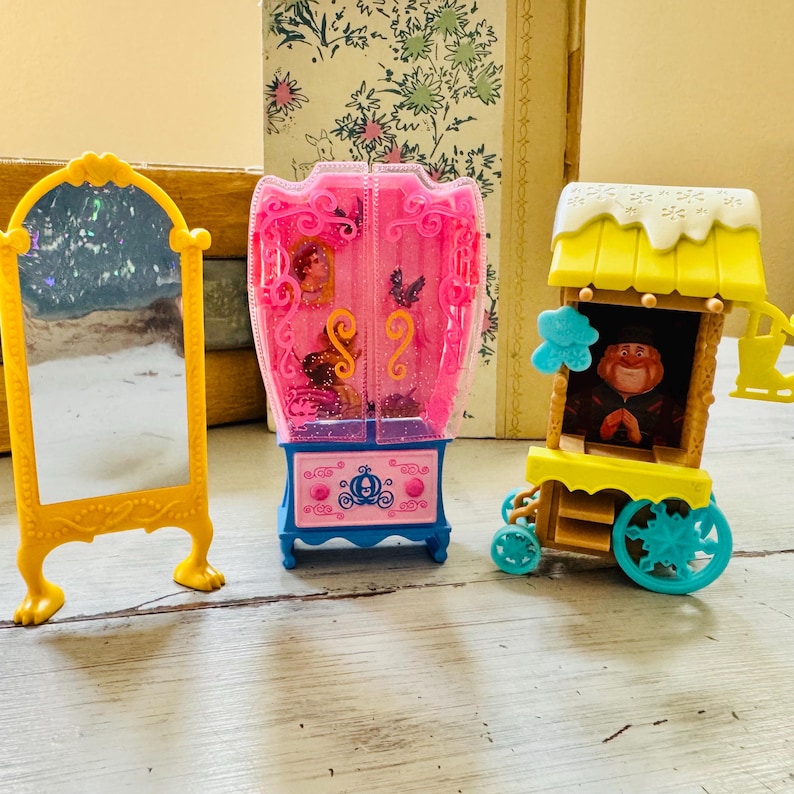 May include: A yellow toy mirror, a pink and blue toy wardrobe, and a yellow and blue toy cart are arranged on a light-colored surface. The mirror has a decorative frame. The wardrobe has a pink frame and blue base. The cart has a character inside.