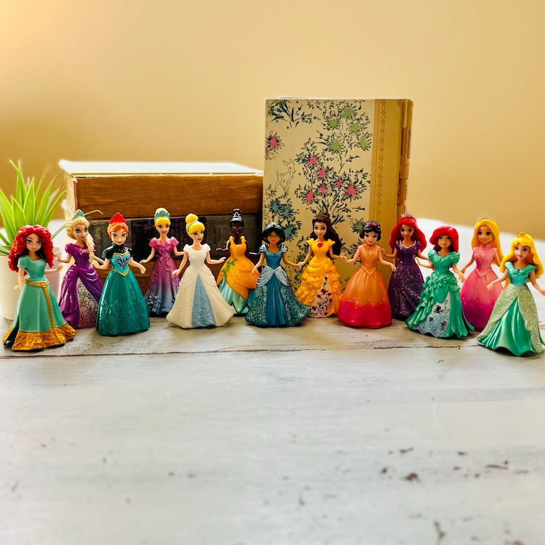 May include: A collection of miniature princess figurines in various colorful gowns. The figurines are arranged in a row, showcasing a variety of dresses in shades of green, blue, pink, yellow, and purple. A wooden box and a book with floral patterns are in the background.