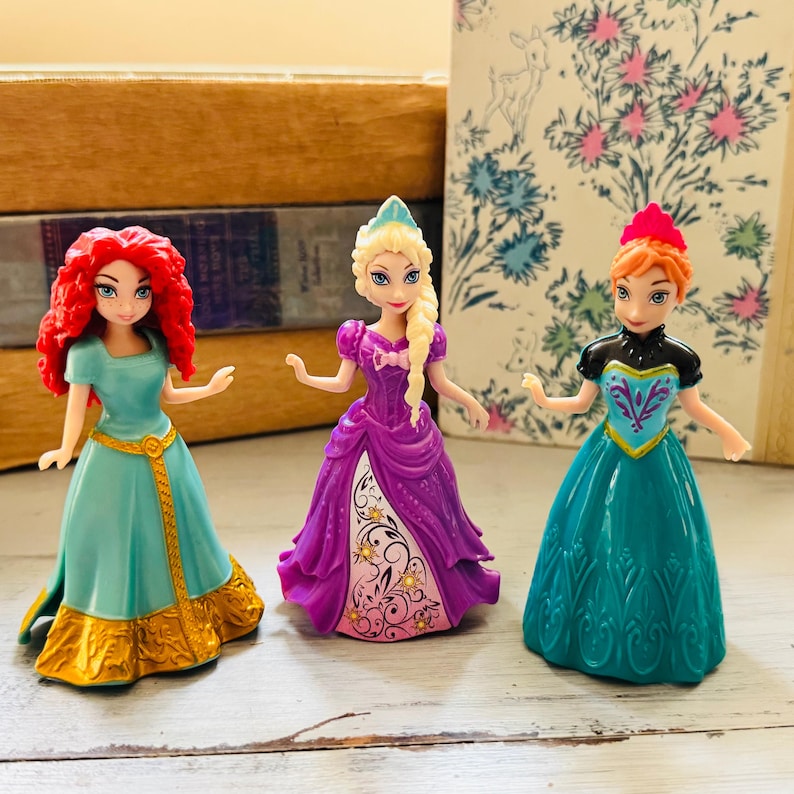 May include: Three miniature princess figurines. One has red hair and a teal dress with gold accents. Another has blonde hair and a purple dress. The third has red hair and a teal dress with a black top.