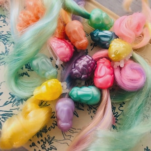 Könnte beinhalten: Eine Sammlung bunter Haaraccessoires aus Kunststoff in verschiedenen Formen und Farbtönen, darunter Rosa, Gelb, Orange und Türkis. Strähnen pastellfarbenen Haares sind mit den Accessoires verwoben und erzeugen eine verspielte und skurrile Ästhetik.