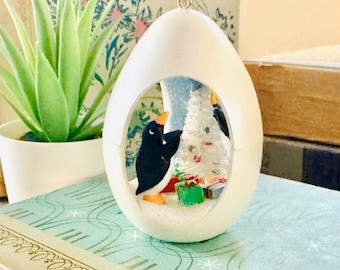 May include: A white egg-shaped Christmas ornament featuring a penguin figurine inside, next to a small Christmas tree. The penguin is black and white with an orange beak and feet. Small wrapped gifts are at the base of the tree. The ornament has a gold loop.