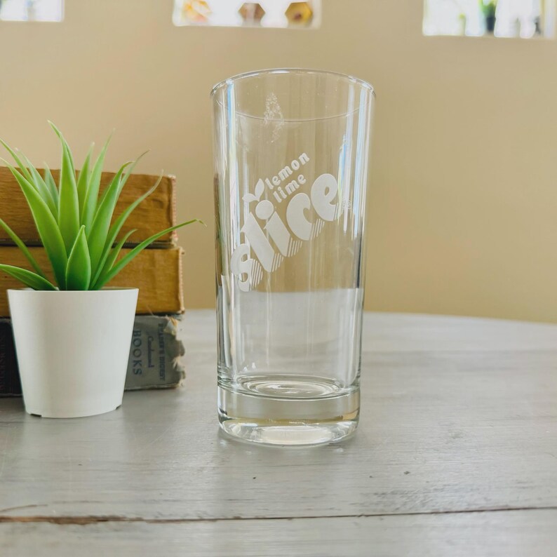 Pu&ograve; includere: Un bicchiere di vetro trasparente con le parole "lemon lime Slice" in un carattere bianco stilizzato. Il bicchiere &egrave; cilindrico e poggia su una superficie chiara. Una piccola pianta in vaso e libri sono sullo sfondo.