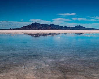 May include: A panoramic view of a vast, blue body of water with a mountain range in the distance. The water is calm and reflects the sky and mountains. The sky is a clear blue with white clouds.