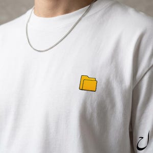 以下が含まれることがあります： 胸に小さな黄色のフォルダのグラフィックが付いた白いTシャツ。Tシャツはクルーネックと半袖です。シルバーのチェーンネックレスが見えます。袖にはアラビア語のスクリプトデザインがあります。