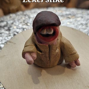 Könnte beinhalten: Eine skurrile Skulptur einer Kreatur mit braunem Hut, beigefarbenem Mantel und rosa Händen. Die Kreatur hat einen großen, offenen Mund mit sichtbaren Zähnen und steht auf einem Holzrund. Der Text "Diddy Eckerslike" steht darüber.