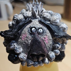 Op de afbeelding: Een grillige, handgemaakte sculptuur van een hondengezicht. De sculptuur heeft een zwarte neus, zwarte oren en een grijs en wit getextureerde vacht. De hond heeft grote, verbaasde ogen en roze wangen. De sculptuur is waarschijnlijk van klei.