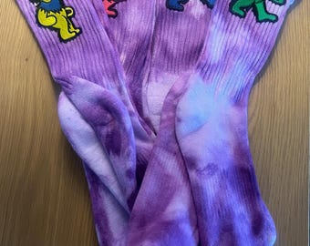 以下が含まれることがあります： 異なる色の刺繍されたクマが各靴下にある、紫色のタイダイ染めの靴下4足セット。クマは黄色、赤、青、緑です。