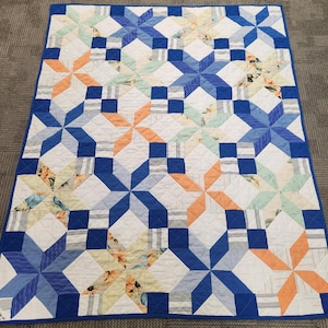 May include: A rectangular quilt featuring a repeating star pattern in shades of blue, peach, and white. The design incorporates solid blue squares and patterned fabric blocks with floral and striped motifs, finished with a solid blue border.