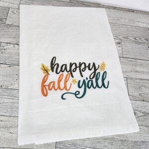 May include: White kitchen towel with embroidered text "happy fall y'all" in black, orange, and green with stylized leaves.