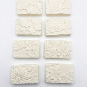 May include: Eight rectangular, off-white soaps with floral and leaf designs. Each soap has a different embossed pattern, including daisies, leaves, and intricate lace-like textures. The soaps are arranged in a grid pattern against a white background.