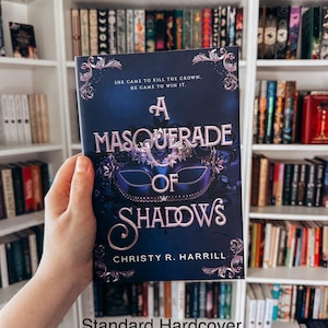 May include: A hardcover book titled "A Masquerade of Shadows" by Christy R. Harrill. The cover is dark blue with a masquerade mask design and ornate floral accents. The text "She came to kill the crown. He came to win it." is also on the cover.