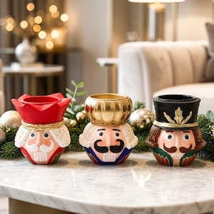 May include: Three decorative nutcracker-themed candle holders arranged in a row. The left holder features a red crown, the centre a gold crown, and the right a black top hat. Each piece displays a painted face with a moustache and white hair.
