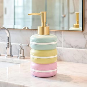 May include: A decorative soap dispenser in the shape of stacked macarons. The dispenser has a gold pump and is made up of light blue, yellow, and pink sections with white detailing. It sits on a marble surface.