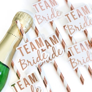 May include: A set of six rose gold and white striped straws with white flags that say "Team Bride" in rose gold lettering.