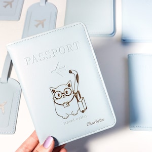 May include: Light blue passport holder with a cat wearing glasses and holding a suitcase. The holder has the word "PASSPORT" and "travel wallet" printed on it. The name "Charlotte" is also printed on the holder.
