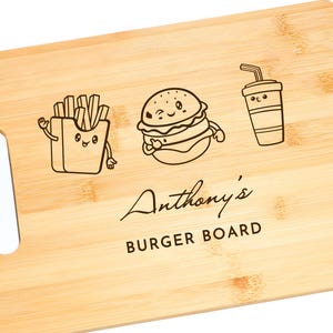Op de afbeelding: Een lichtbruin houten snijplank met een handvat. De plank heeft een cartoon hamburger, frietjes en een drankje met een rietje erop. De tekst "Anthony's Burger Board" is in de plank gegraveerd.