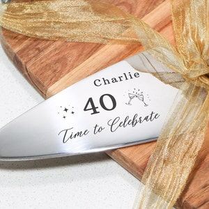 May include: A silver serving knife with a gold ribbon tied around it. The knife is engraved with the text "Charlie 40 Time to Celebrate" and a small image of two champagne glasses.