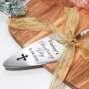 Può includere: Un servizio per torta in argento con un nastro dorato legato attorno al manico. Il servizio è inciso con il testo "Finley's Christening Day 10.08.2024" e una croce.