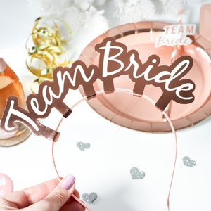 May include: Rose gold headband with the words "Team Bride" in white lettering. The headband is on a pink plate with a rose gold rim. There are two smaller rose gold "Team Bride" signs on the table.