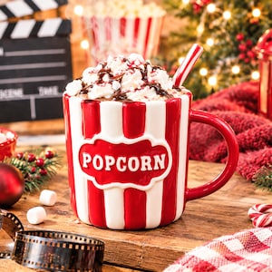 Könnte beinhalten: Ein Keramikbecher mit vertikalen roten und weißen Streifen und einem erhabenen roten Etikett mit dem Wort POPCORN in weißen Buchstaben. Der Becher ist mit einem Getränk gefüllt, das mit Schlagsahne, Schokoladensauce und einem Strohhalm garniert ist.