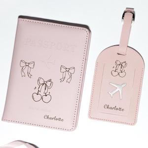 May include: Pink passport holder and matching luggage tag with a silver airplane, cherries, and bows. The passport holder says "PASSPORT" and "Charlotte". The luggage tag says "Charlotte".