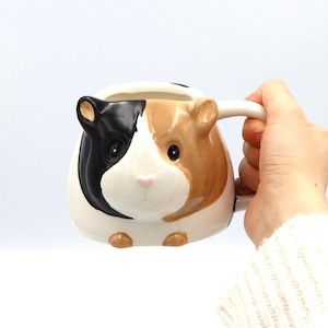 May include: A ceramic guinea pig mug with a white handle and a detailed design. The mug features a black, brown, and white guinea pig face with small ears and a pink nose. The mug is held by a person wearing a white sweater.