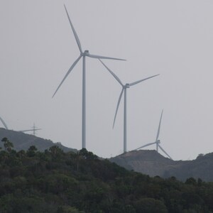 May include: A landscape view of wind turbines on a hillside. The turbines are white and have three blades each. The sky is overcast, and the foreground is filled with green trees and vegetation.
