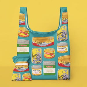 Könnte beinhalten: Eine türkisfarbene, wiederverwendbare Einkaufstasche mit einem sich wiederholenden Muster aus bunten Konserven. Die Dosen zeigen Bilder von Fisch, Erdbeeren und anderen Lebensmitteln mit Text in mehreren Sprachen. Eine passende kleine Tasche ist befestigt.