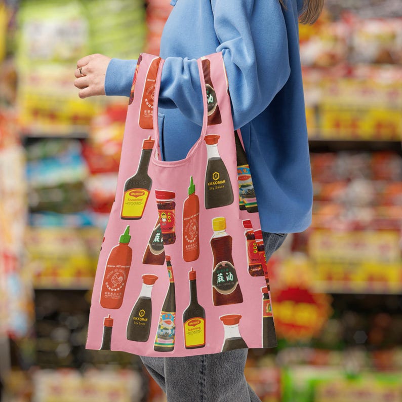 May include: A pink reusable shopping bag featuring a repeating pattern of various Asian sauces and condiments. The bag is held by a person in a blue sweatshirt in a grocery store setting. The bag's design includes soy sauce, chili sauce, and other bottled sauces.