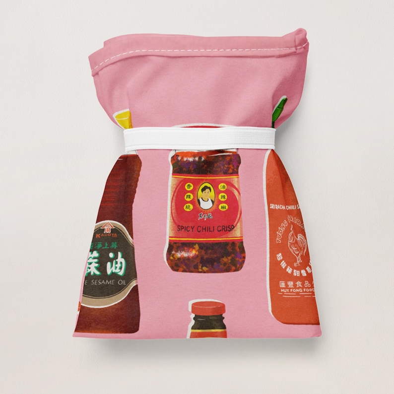 May include: A pink fabric bag featuring illustrations of Asian condiments. The design includes bottles of sesame oil, spicy chili crisp, and sriracha chili sauce. The bag is tied with a white band.
