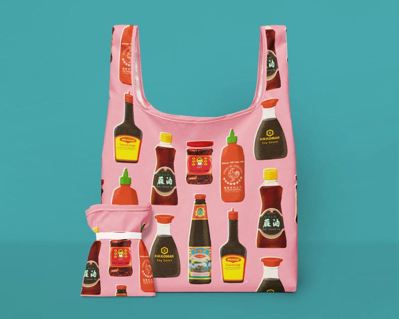 May include: Pink reusable shopping bag with a pattern of various Asian sauces and condiments. The bag includes a matching pouch. The sauces are in bottles and jars of different shapes and sizes, with colorful labels.