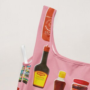 May include: Pink tank top featuring a pattern of various condiment bottles, including Sriracha, Maggi seasoning, and other sauces. The top has a white strap and a pink trim.
