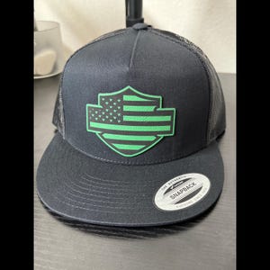 May include: Black snapback hat with a green and white American flag emblem. The hat features a mesh back and a curved brim. The emblem is a shield shape with stars and stripes.