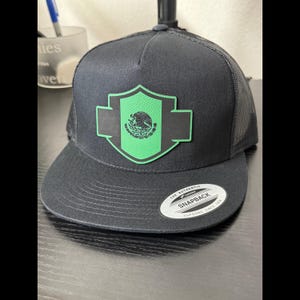 May include: Black trucker hat with a flat brim and mesh back. Features a green and black emblem with the Mexican coat of arms. The hat has a snapback closure and a white tag that reads "The Authentic Snapback".