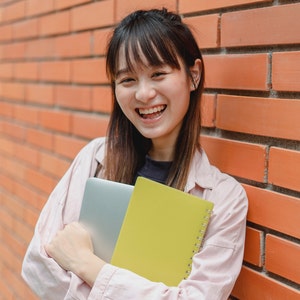 May include: A young person with long brown hair is smiling while holding a laptop and a yellow notebook. They are standing in front of a brick wall.