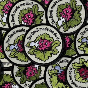 May include: Circular embroidered patch with the text "The Devil made me do it." The patch features a pink devil character inside a green lettuce-like design, with a white background and black border.
