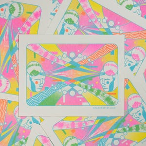 May include: A stack of art prints with a vibrant, abstract design. The prints feature a central image with a pink, yellow, and blue color scheme, with geometric shapes and stylized human figures. The text "STARKEEP STUDIO" is visible.