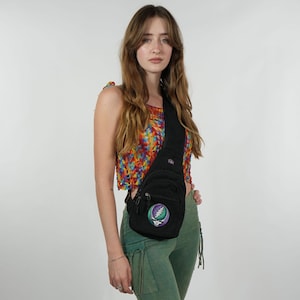 May include: Black sling bag with a zippered main compartment and a round patch featuring a skull and lightning bolt design in purple, green, and white. The bag is worn across the body. The model is wearing a colorful crochet top and green pants.