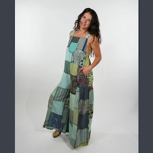 May include: A patchwork jumpsuit in shades of blue, green, and gray. The wide-leg jumpsuit features a variety of patterned fabric squares sewn together. It has a bib top with adjustable straps and pockets. The model is wearing brown shoes.